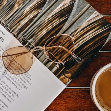 Close-up of stylish sunglasses resting on a magazine beside a cup of coffee on a wooden table.