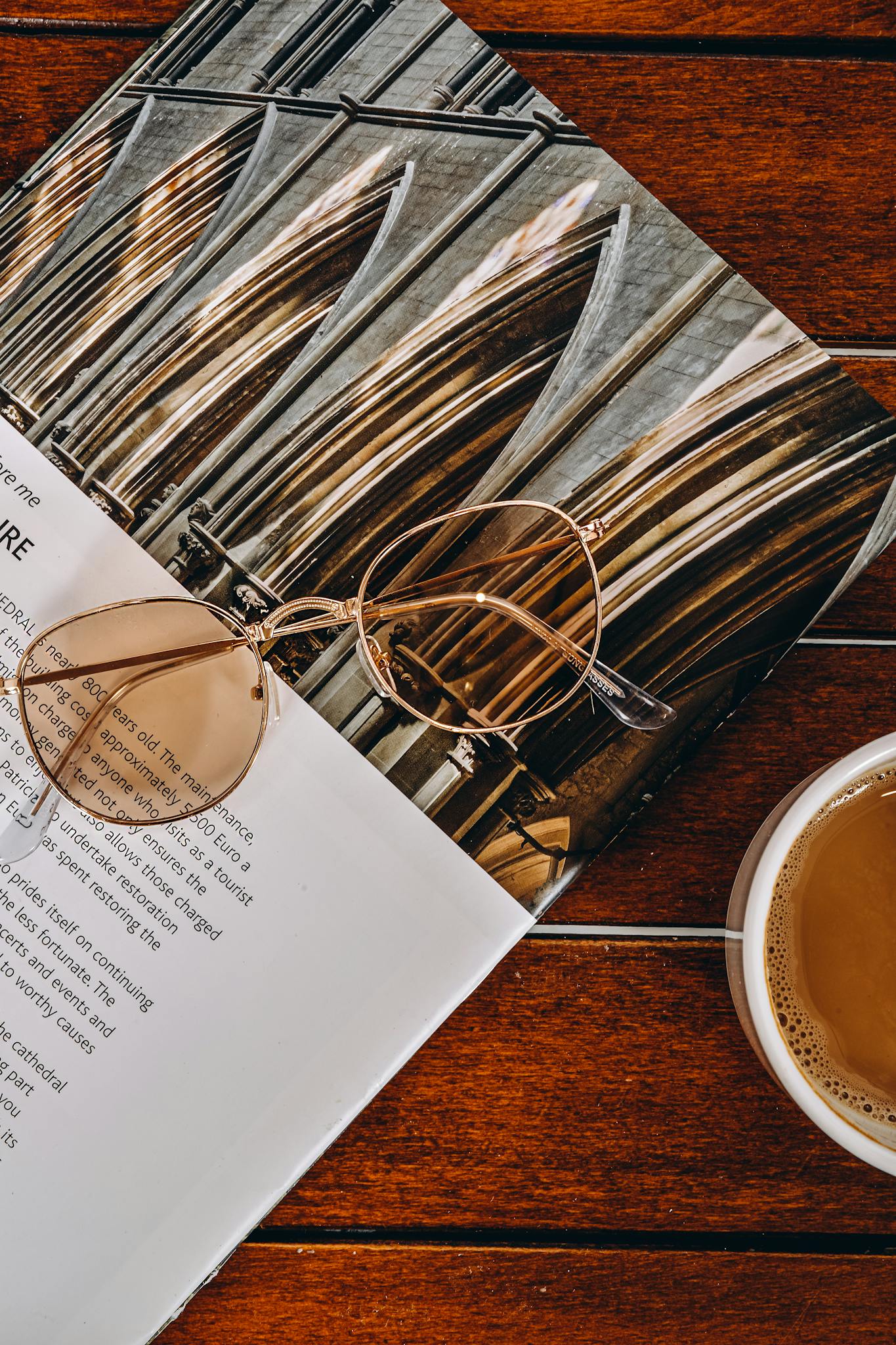 Close-up of stylish sunglasses resting on a magazine beside a cup of coffee on a wooden table.