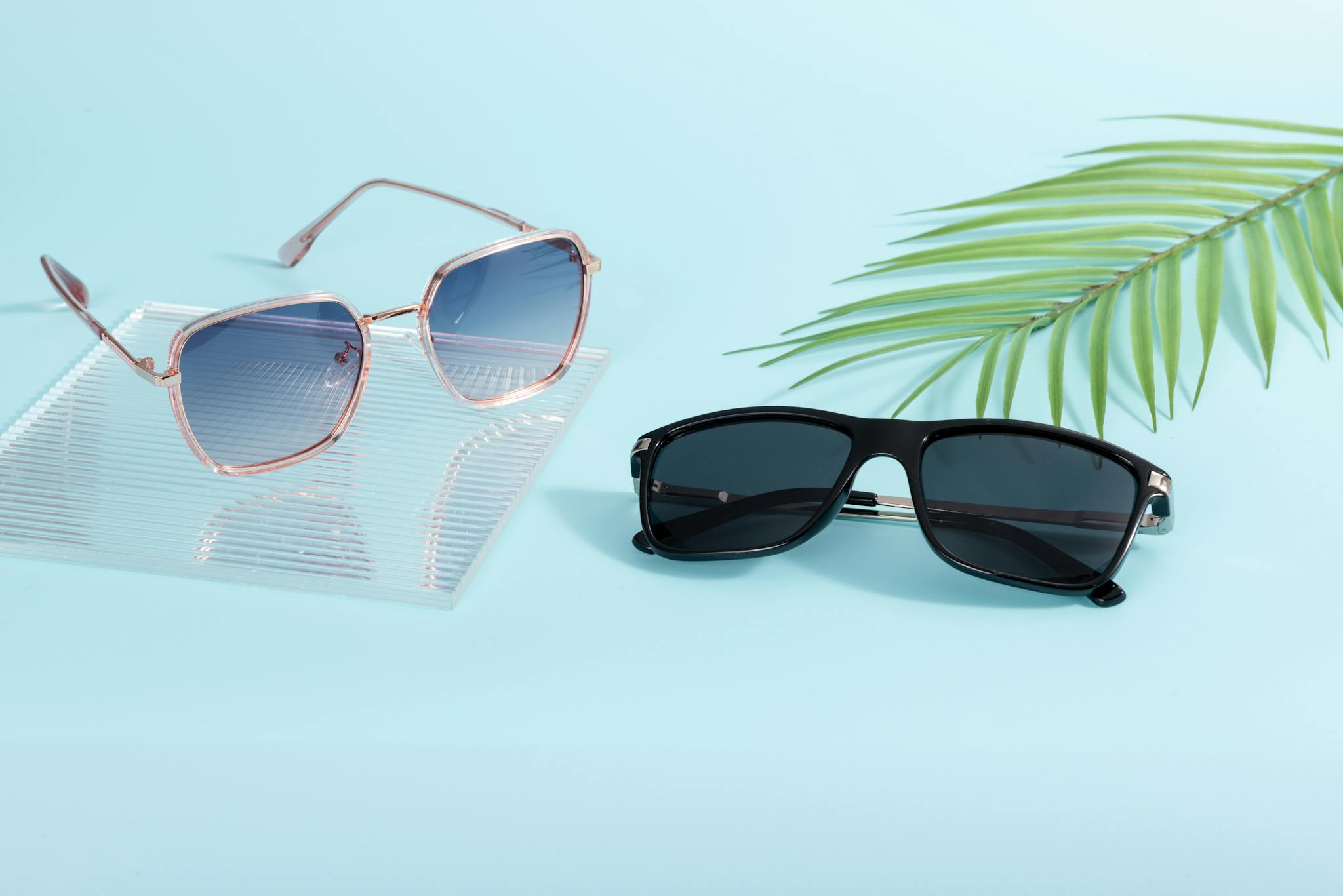Two pairs of trendy sunglasses on a pastel surface with palm leaf decor.